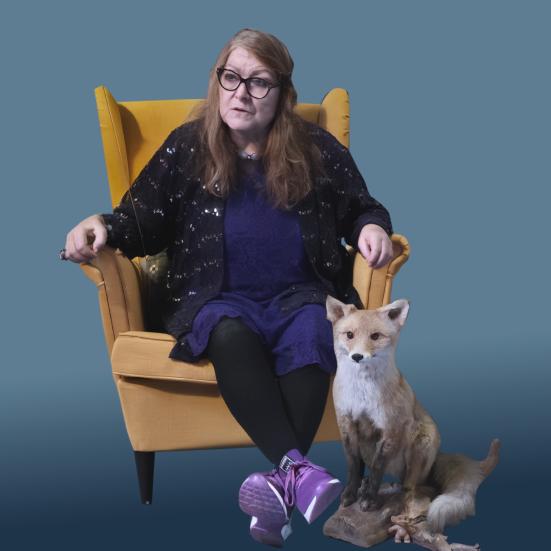 Dame sidder i en gul stol, ved siden af hende står en udstoppe ræv.