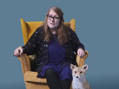 Dame sidder i en gul stol, ved siden af hende står en udstoppe ræv.
